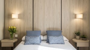 Un dormitorio con cabecera de madera, dos mesitas de noche con lámparas, una cama con almohadas estampadas, un florero con flores y una planta pequeña en la mesita de noche.