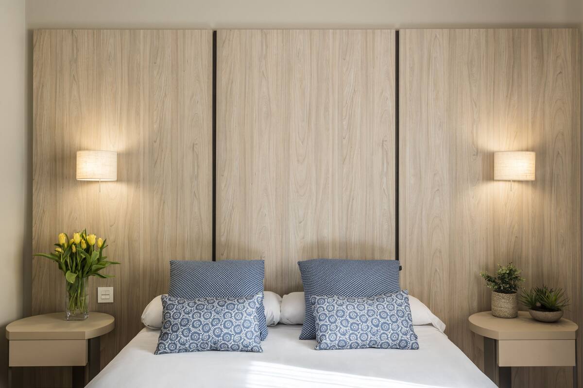 Ein Schlafzimmer mit einem Holz-Kopfteil, zwei Nachttischen mit Lampen, einem Bett mit gemusterten Kissen, einer Vase mit Blumen und einer kleinen Pflanze auf dem Nachttisch.