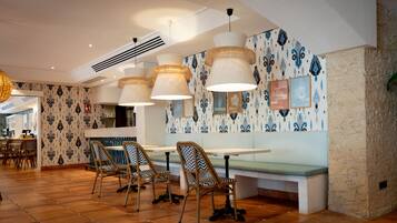 Interior de restaurante con techo alto, papel tapiz estampado y pared de acento de piedra. Se observan mesas y sillas dispuestas para comer.