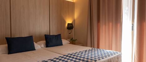 A double bed with a geometric patterned bedspread, two pillows, a bedside table with a plant, and a wall-mounted light fixture.