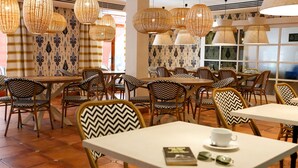 Un restaurante con mesas y sillas redondas, lámparas colgantes de mimbre, papel tapiz estampado y piso de baldosas.