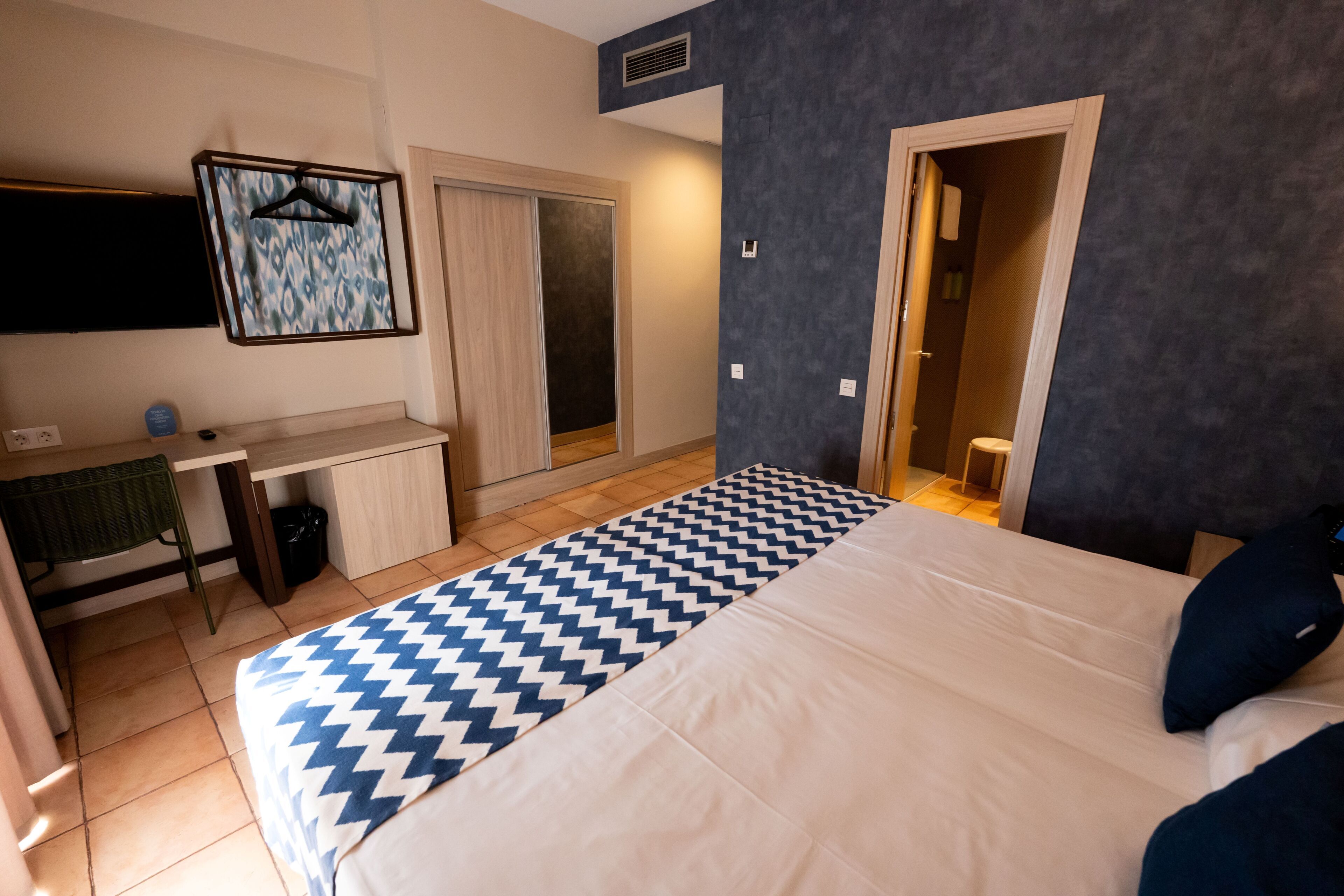 A hotel room with a bed, a desk, a chair, a wardrobe, and a bathroom visible through an open door.