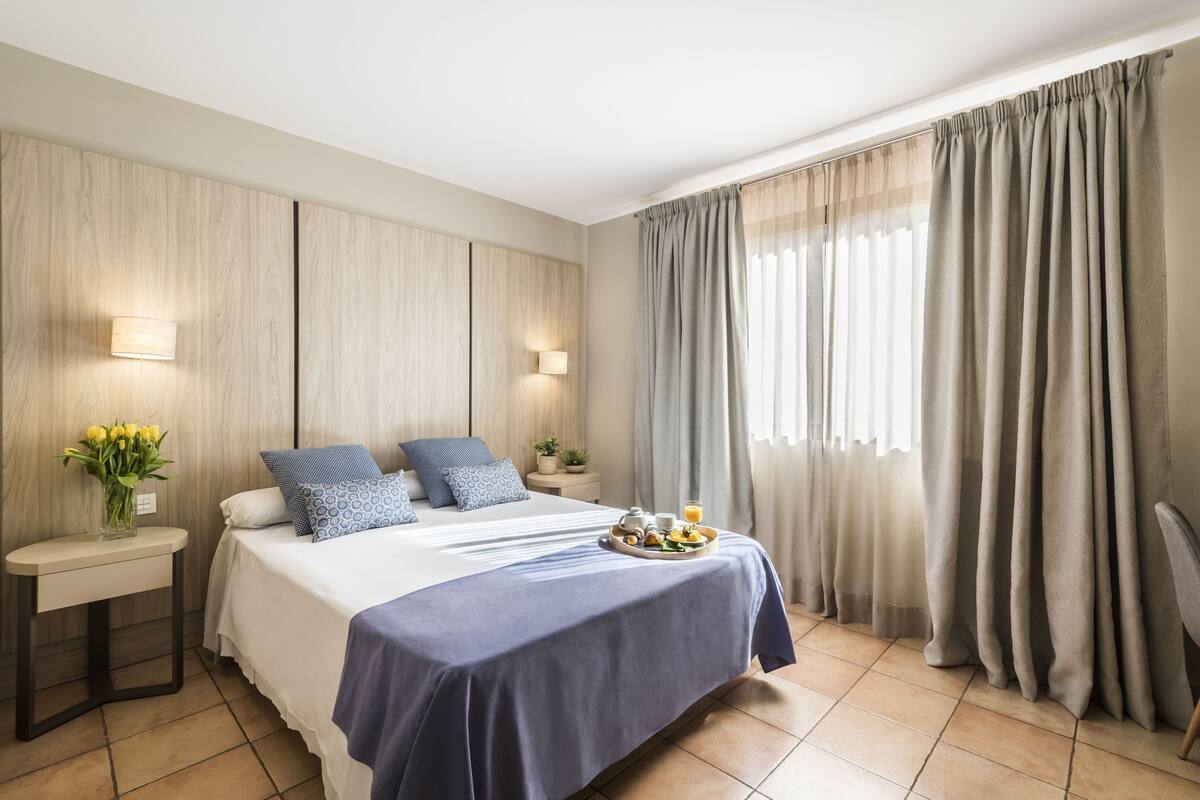 Ein modernes Hotelzimmer mit einem großen Bett, zwei Nachttischen, einem Beistelltisch, einer Vase mit Blumen und einem Frühstücksbrettchen auf dem Bett.