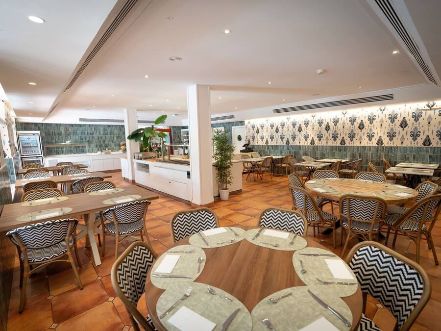 En restaurant med runde borde, stole med mønstrede hynder og en flisebeklædt væg.