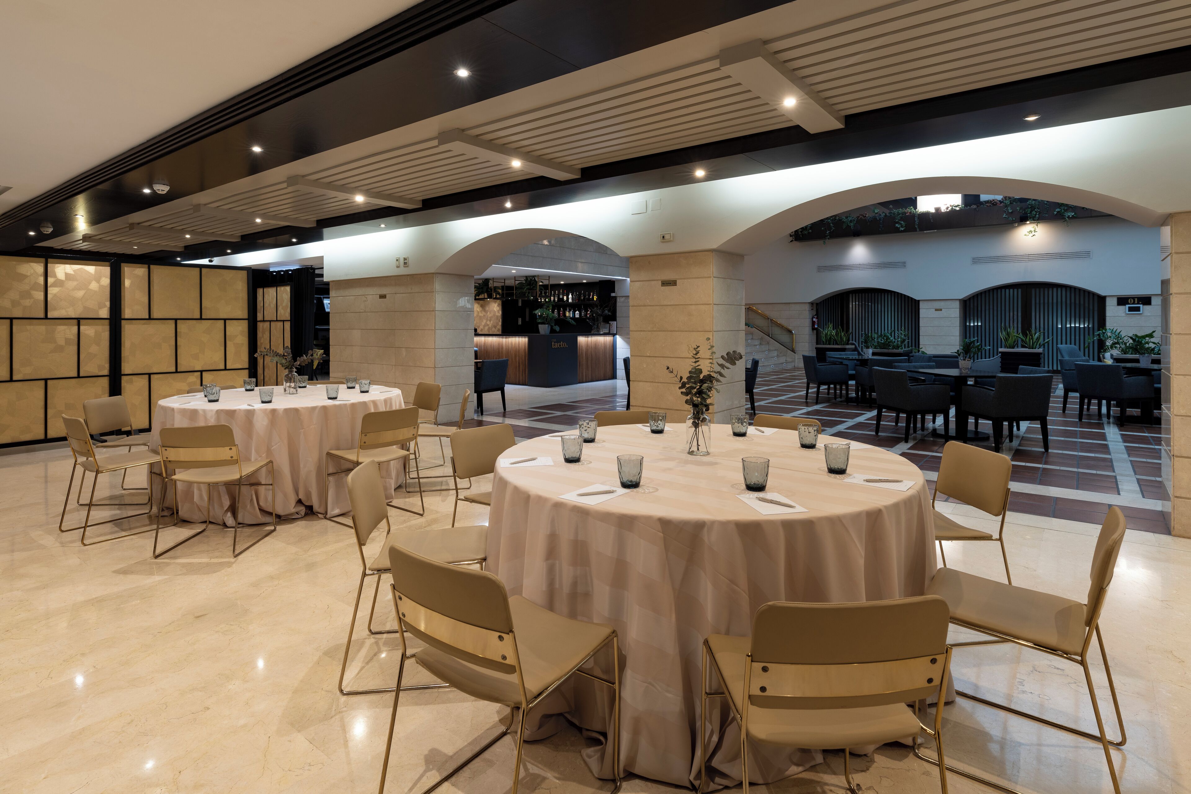 Un amplio hall de hotel con varias mesas y sillas redondas, una zona de bar y una escalera.