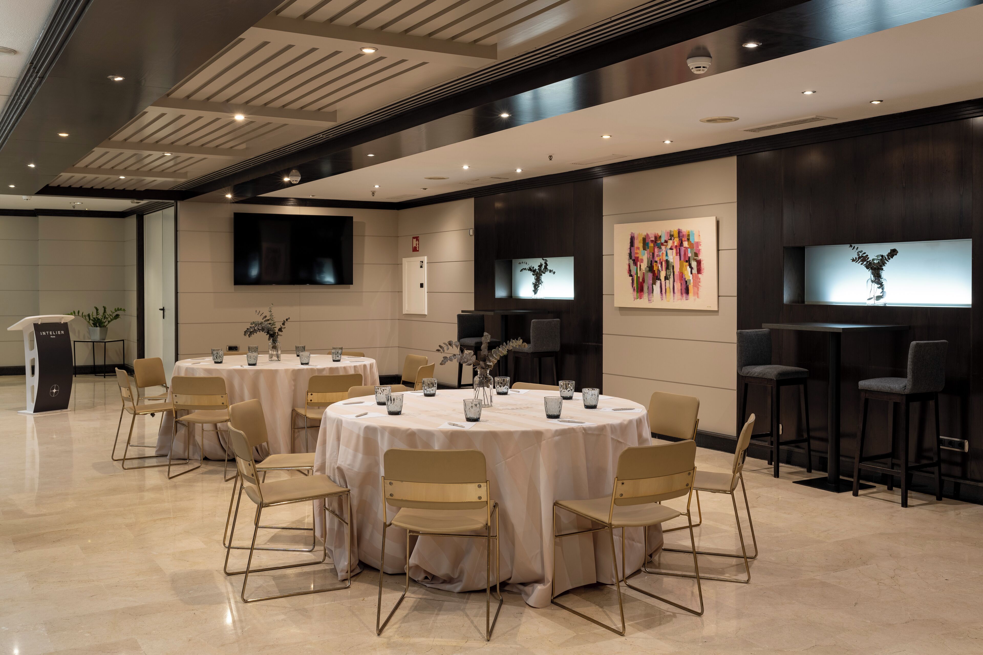 A modern conference room with round tables, beige chairs, and a flat-screen TV on the wall.