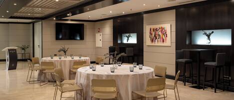 A modern conference room with round tables, beige chairs, and a flat-screen TV on the wall.
