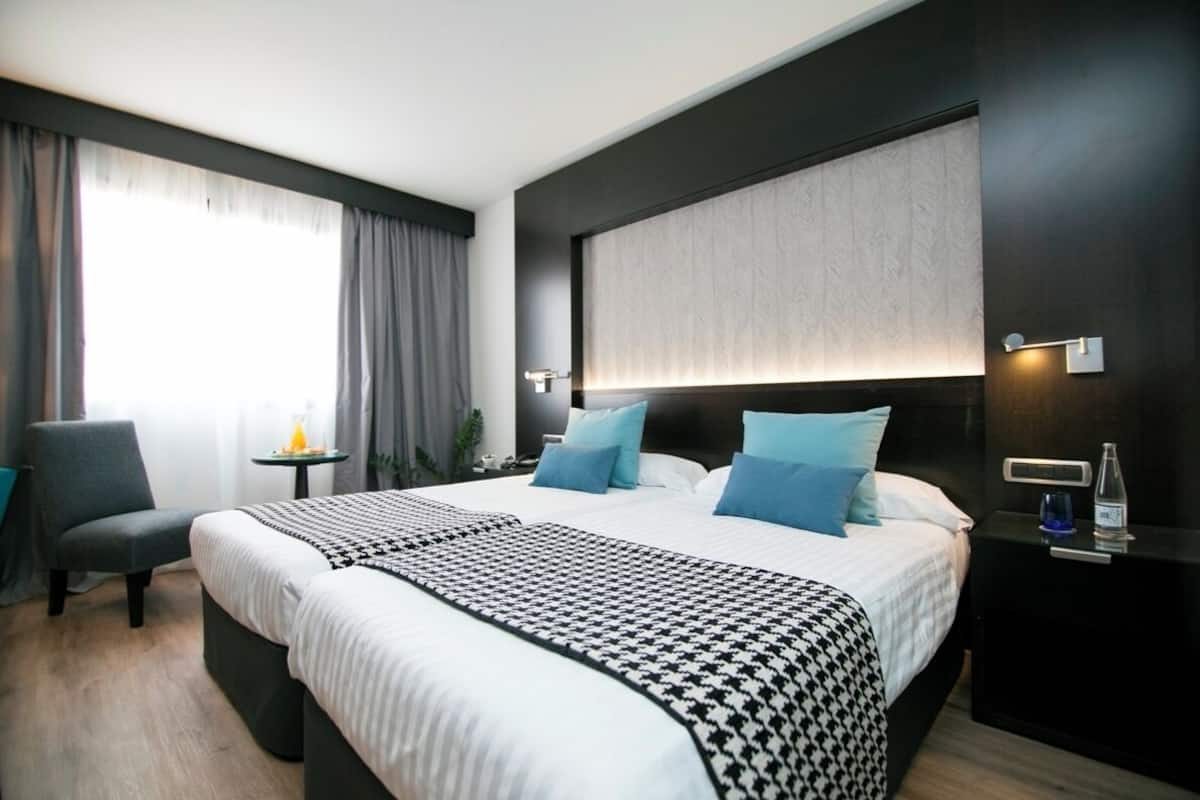 A modern hotel room with a large bed, a bedside table with a lamp, a chair, and a view of the outdoors.