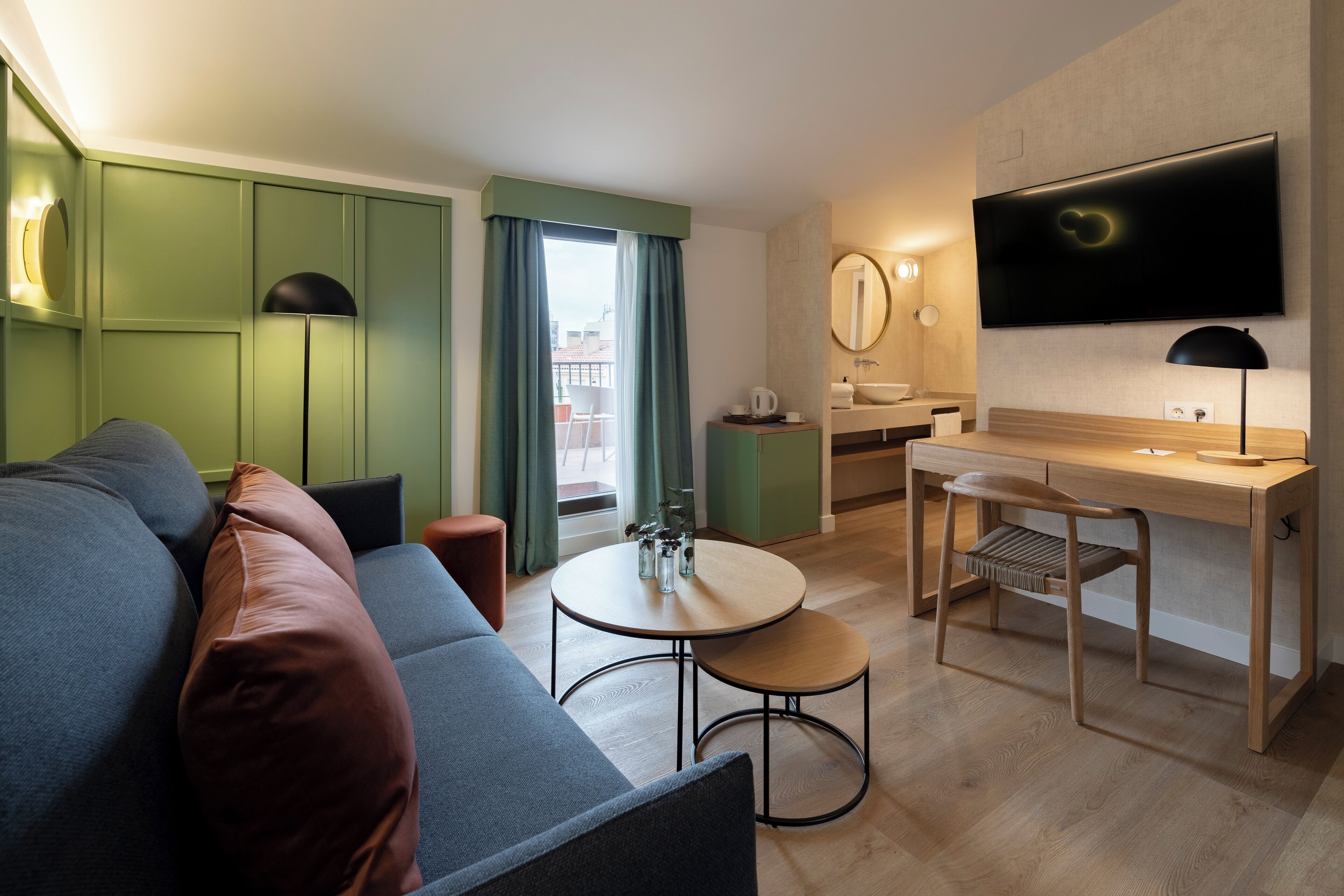 A modern hotel room with a sofa, a small round table, a desk with a chair, a TV mounted on the wall, and a window with curtains.