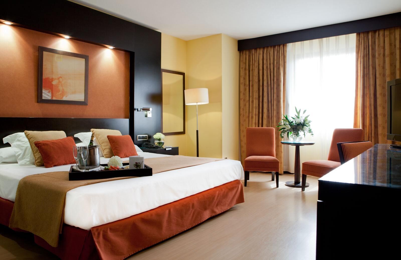 A hotel room with a large bed, a desk, two chairs, a small table with a plant, and a large window with curtains.