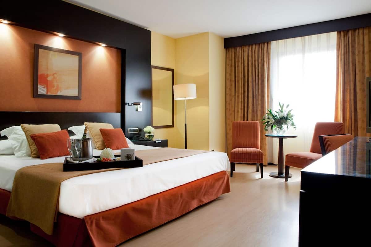 A hotel room with a large bed, a desk, two chairs, a small table with a plant, and a large window with curtains.