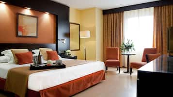A hotel room with a large bed, a desk, two chairs, a small table with a plant, and a large window with curtains.