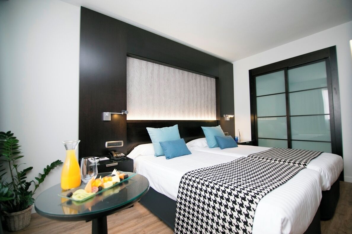 A hotel room with a bed, bedside table with breakfast, a large window, and a potted plant.