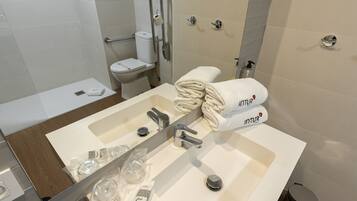 Un baño moderno con un lavamanos blanco, dos grifos, un espejo y un inodoro. Hay toallas y artículos de aseo en el lavamanos.