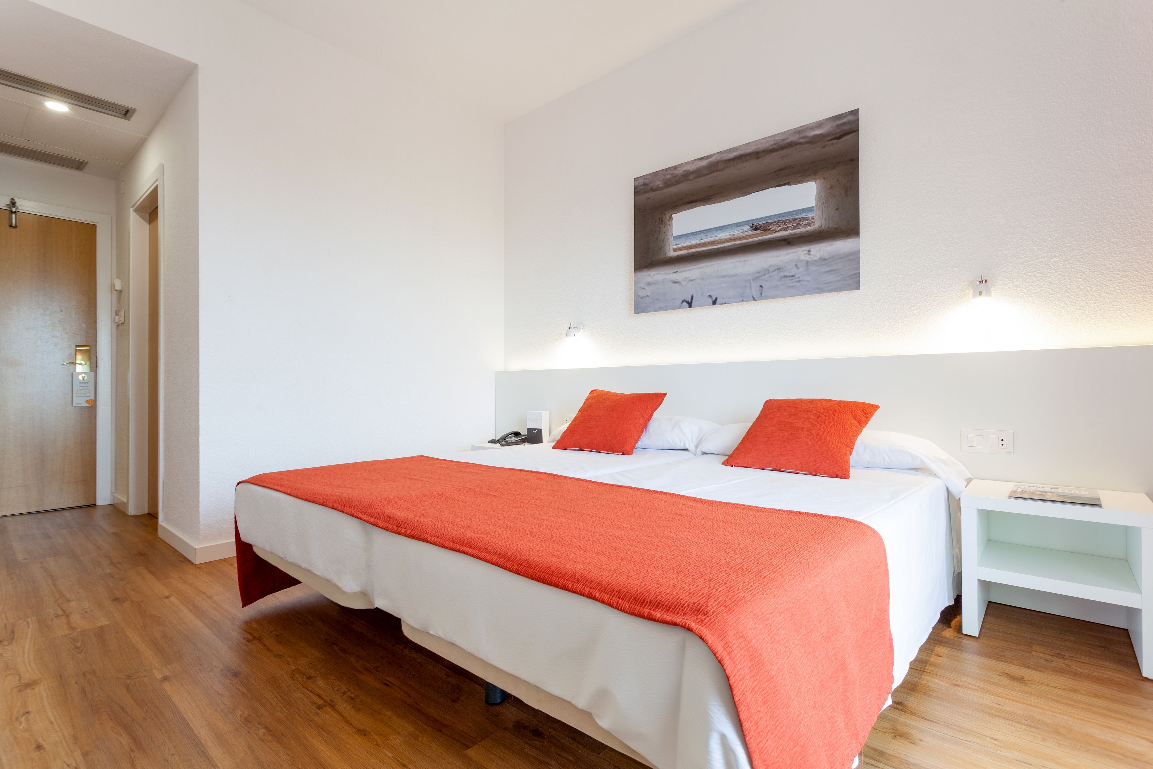Een hotelkamer met een groot bed, wit beddengoed en een rode deken. Er hangt een schilderij aan de muur, en er is een nachtkastje.