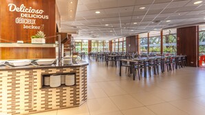 En rummelig kantine med flere borde og stole, et buffetbord med stablede tallerkener, og store vinduer med udsigt til grøn vegetation udenfor.