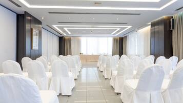 Une salle de conférence avec des rangées de chaises et de tables recouvertes de tissu blanc, un design de plafond moderne et de grandes fenêtres dotées de rideaux.
