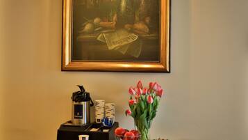 Una estación de café con una cafetera Dallmayr, una taza, un jarrón con tulipanes, un cuenco de fruta y un cuadro en la pared.