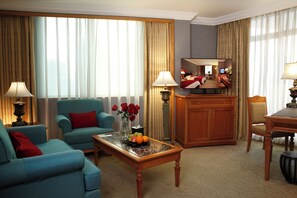 A hotel room with a blue sofa, a wooden coffee table with fruit and flowers, a wooden desk, and a large mirror. - Grand Noble Hotel (Dongguan)