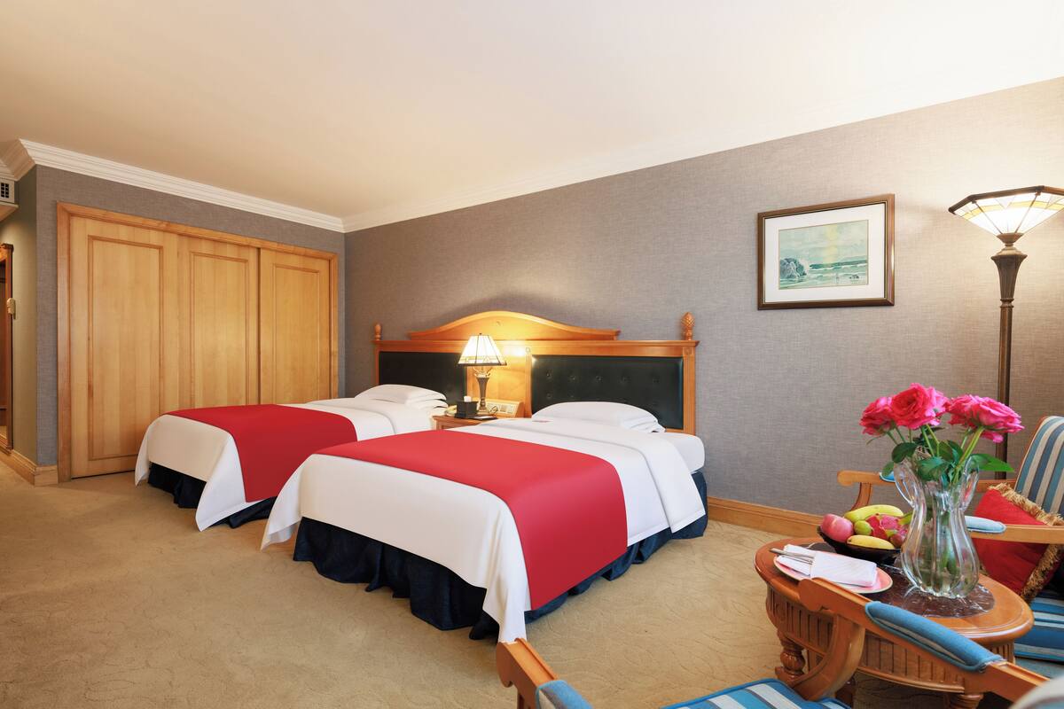 Habitación de hotel con dos camas, una mesita de noche con lámpara, una silla, una pequeña mesa con un jarrón de flores y un cuadro en la pared.