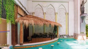 A pool area with a thatched roof seating area, a stone wall with arches, and a statue. - Grand Noble Hotel (Dongguan)