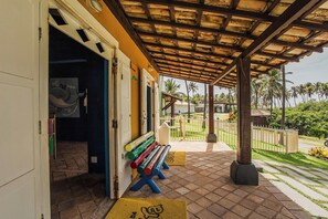 Área externa coberta com um banco, uma porta e vista para palmeiras e um gramado.
