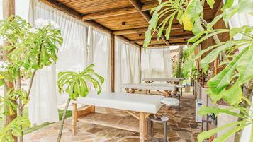 An outdoor covered patio with stone flooring, wooden furniture, and lush green plants.