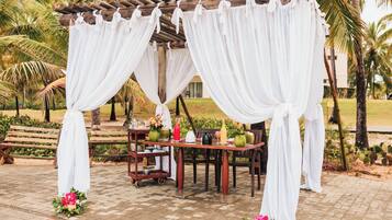 Uma pérgola de madeira com cortinas brancas, uma mesa com um vaso verde e um banco.