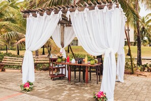 Una pérgola de madera con cortinas blancas, una mesa con un jarrón verde y un banco.