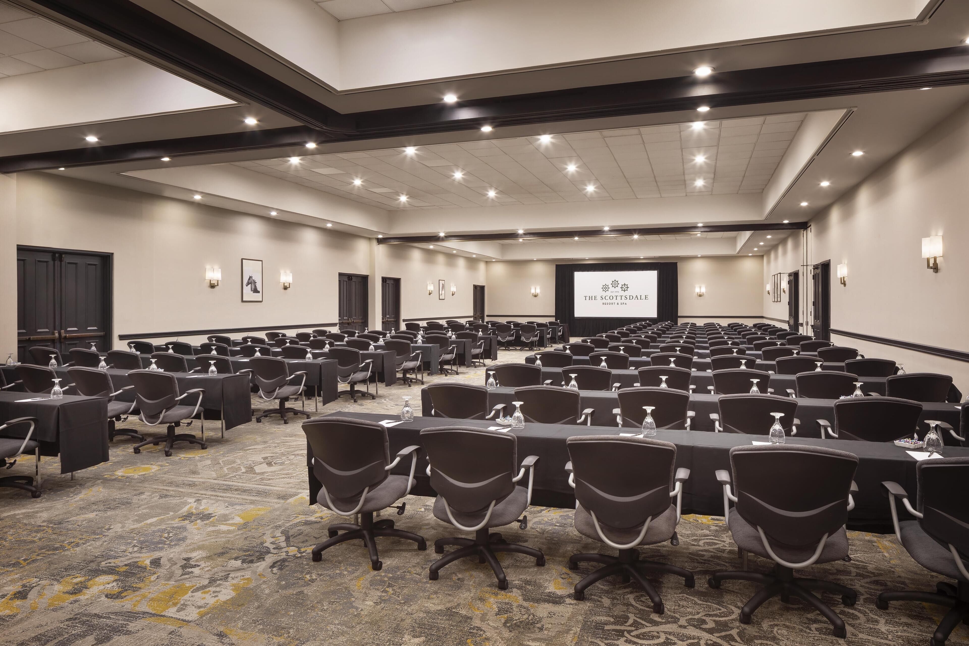 Uma grande sala de conferências com fileiras de mesas e cadeiras, equipada com garrafas de água e uma tela de projetor exibindo "The Scottsdale Resort & Spa".