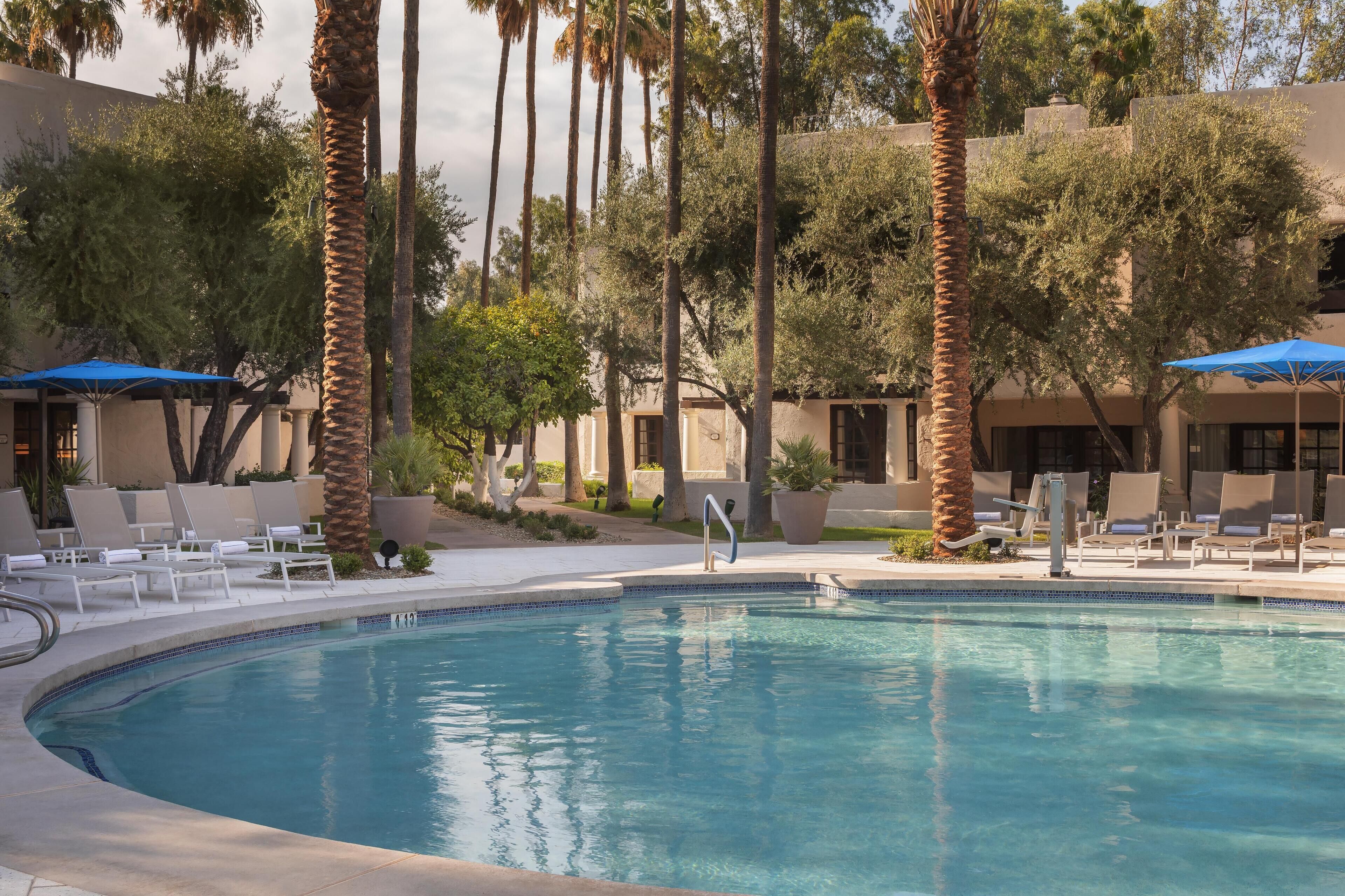 A pool area with lounge chairs and umbrellas, surrounded by palm trees and a building with a beige facade.