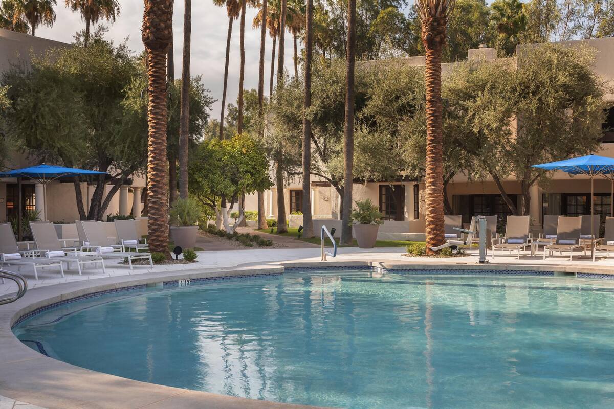 A pool area with lounge chairs and umbrellas, surrounded by palm trees and a building with a beige facade.