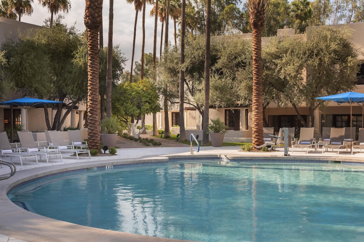 A pool area with lounge chairs and umbrellas, surrounded by palm trees and a building with a beige facade.