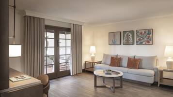 Una sala de estar moderna con un sofá, una mesa de centro y lámparas. Hay un escritorio con una lámpara y una silla. La habitación tiene ventanas grandes con cortinas.