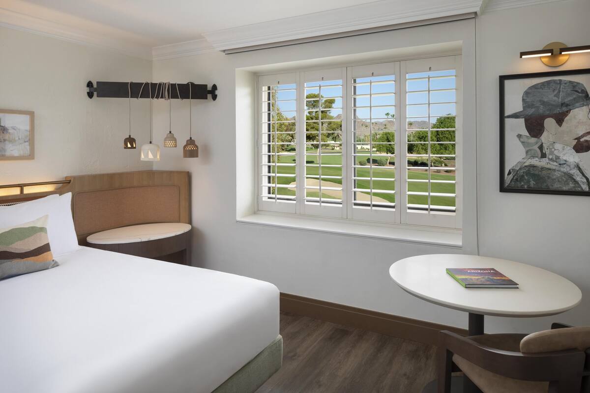 A hotel room with a bed, a small round table, a chair, a window with a view of a golf course, and pendant lights.