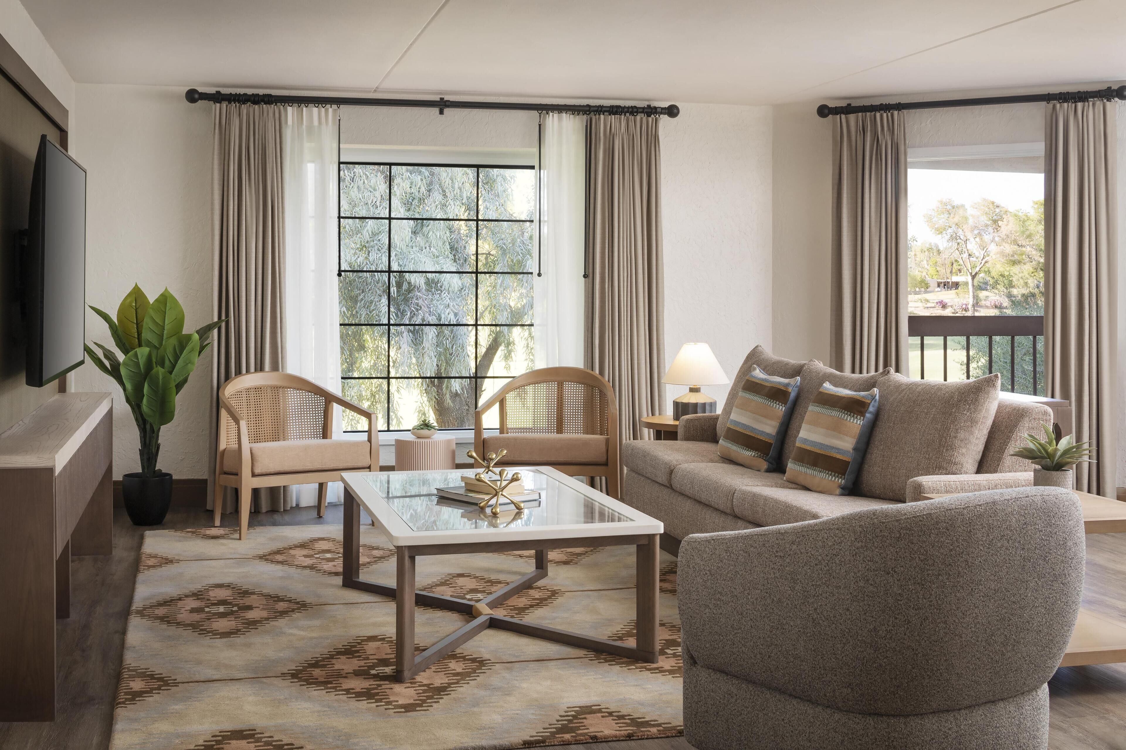 Een woonkamer met een bank, twee fauteuils, een salontafel en een televisie. Er is een groot raam met gordijnen, een kamerplant en uitzicht op groen.