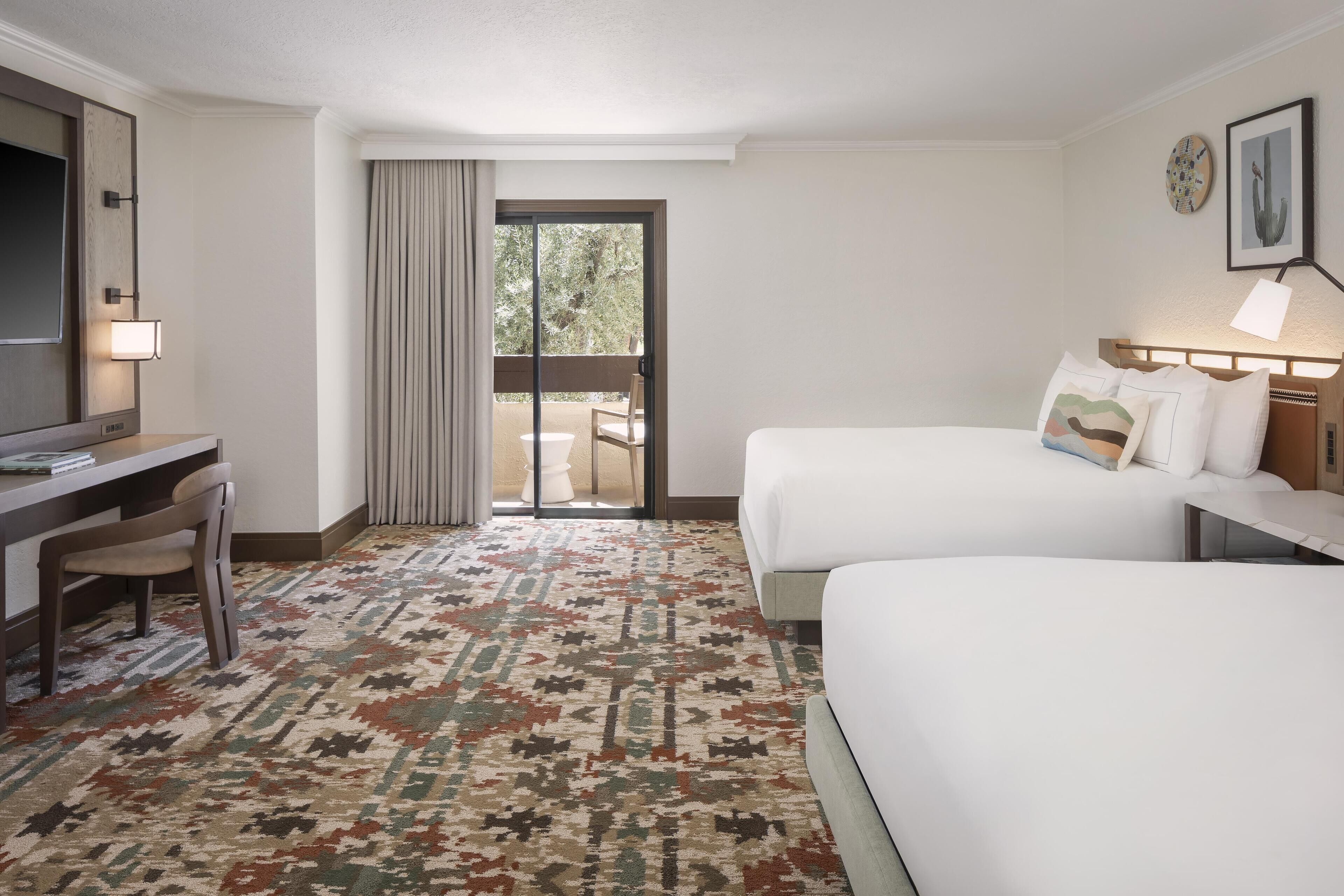A hotel room with two beds, a desk, and a chair. There is a large window with a view of greenery outside.