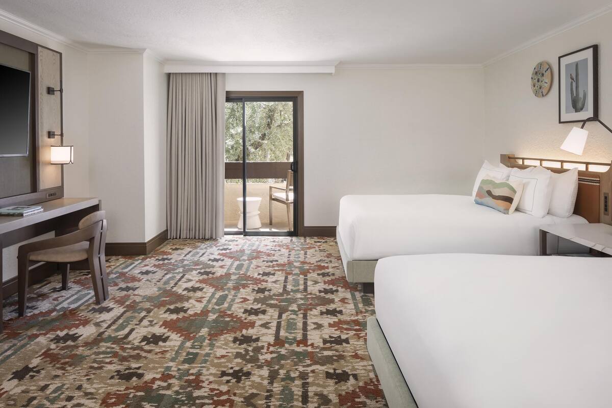 A hotel room with two beds, a desk, and a chair. There is a large window with a view of greenery outside.