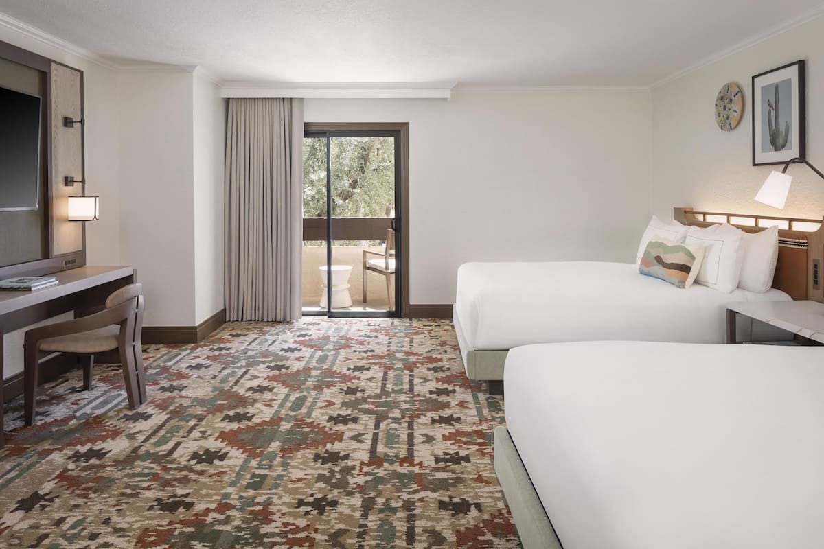 A hotel room with two beds, a desk, and a chair. There is a large window with a view of greenery outside.