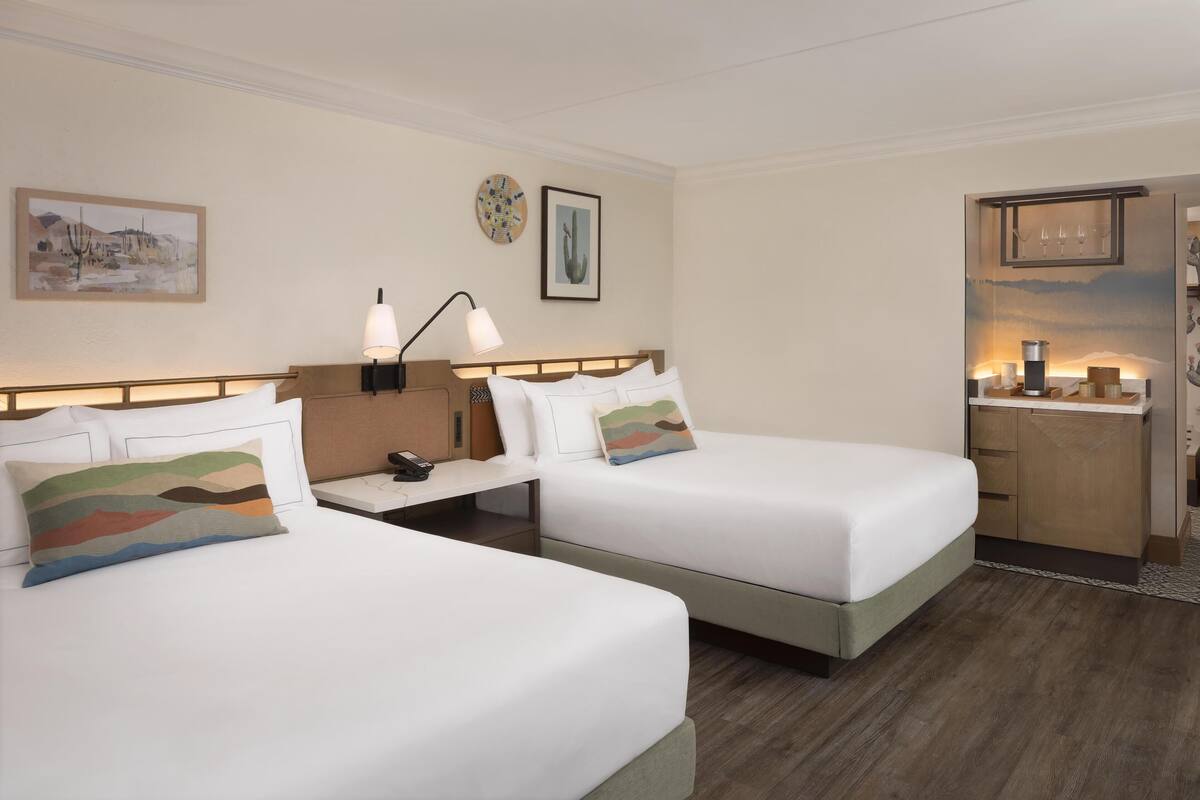 A hotel room with two beds, a bedside table with a lamp, a small desk, and a kitchenette area.
