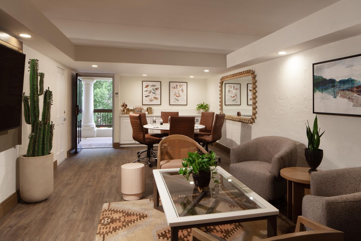 A modern living room with a glass coffee table, a sofa, a dining area with chairs, a large mirror, and a cactus plant.
