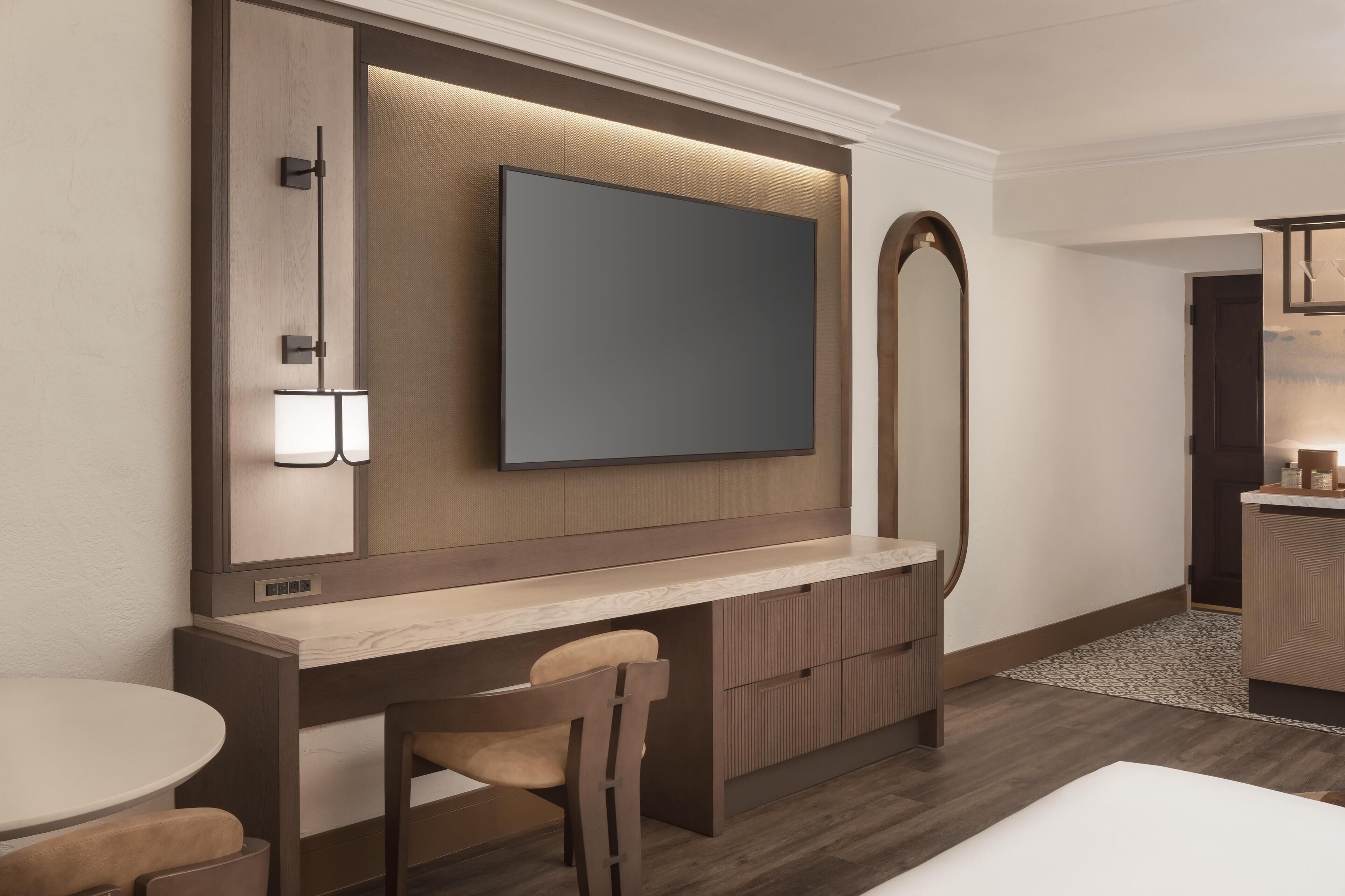 A modern hotel room with a large flat-screen TV mounted on a wooden panel, a small desk with a chair, and a compact bathroom area.