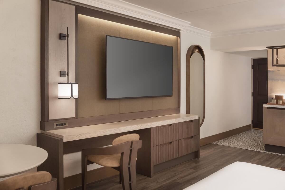 A modern hotel room with a large flat-screen TV mounted on a wooden panel, a small desk with a chair, and a compact bathroom area.