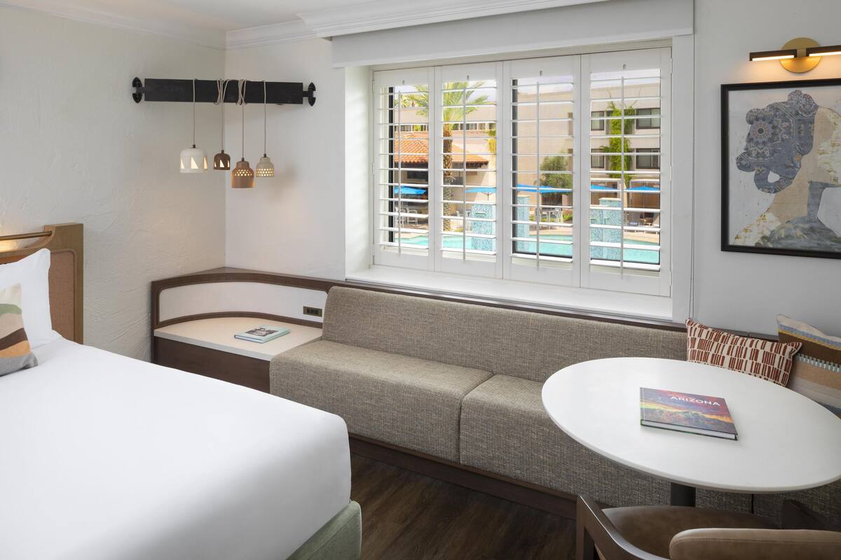 A hotel room with a bed, a sofa, a small table, a window with a view of a pool and buildings, and a framed picture on the wall.