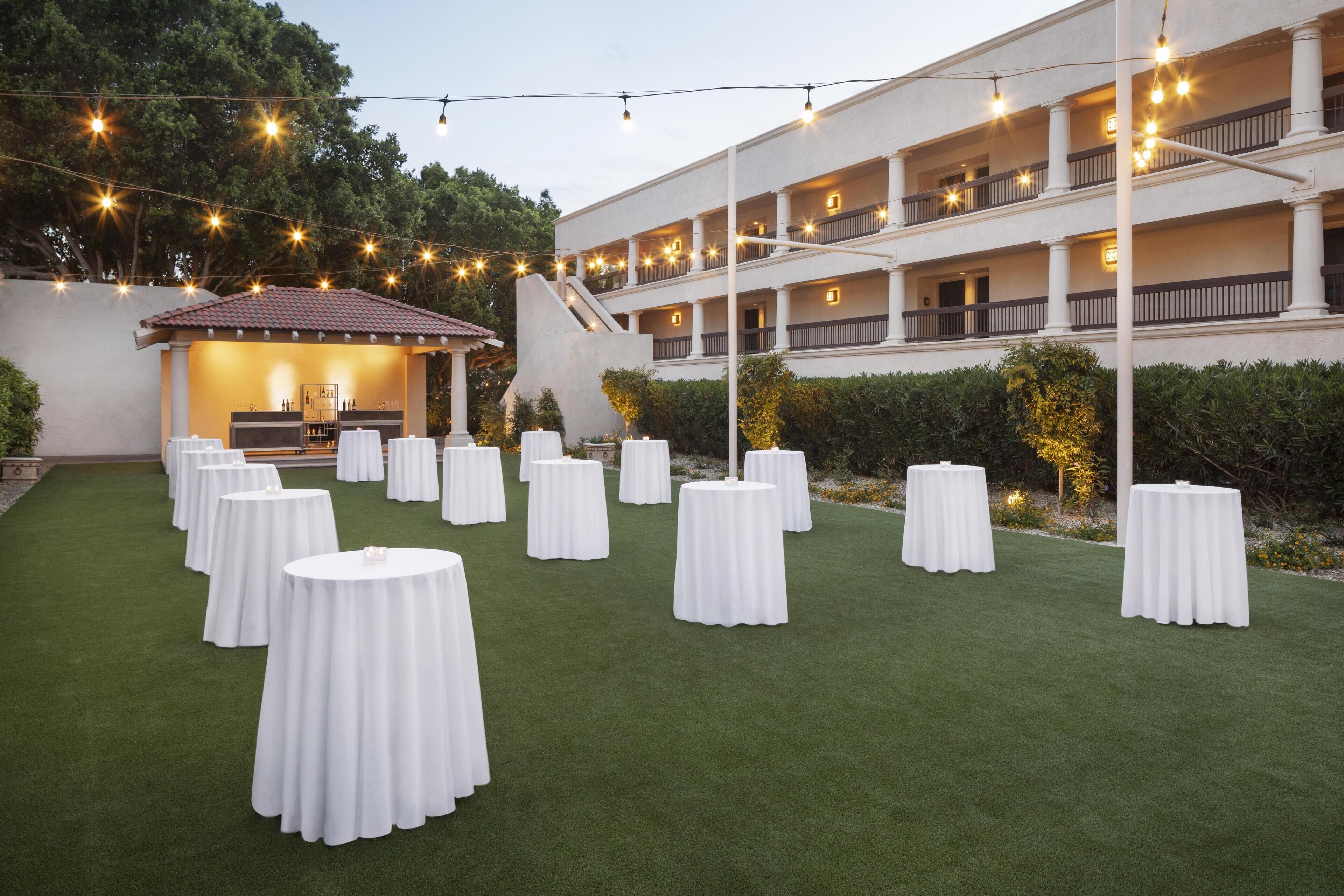 Un espace extérieur aménagé pour les événements, avec plusieurs tables rondes recouvertes de nappes blanches, un espace de repos abrité et des guirlandes lumineuses.