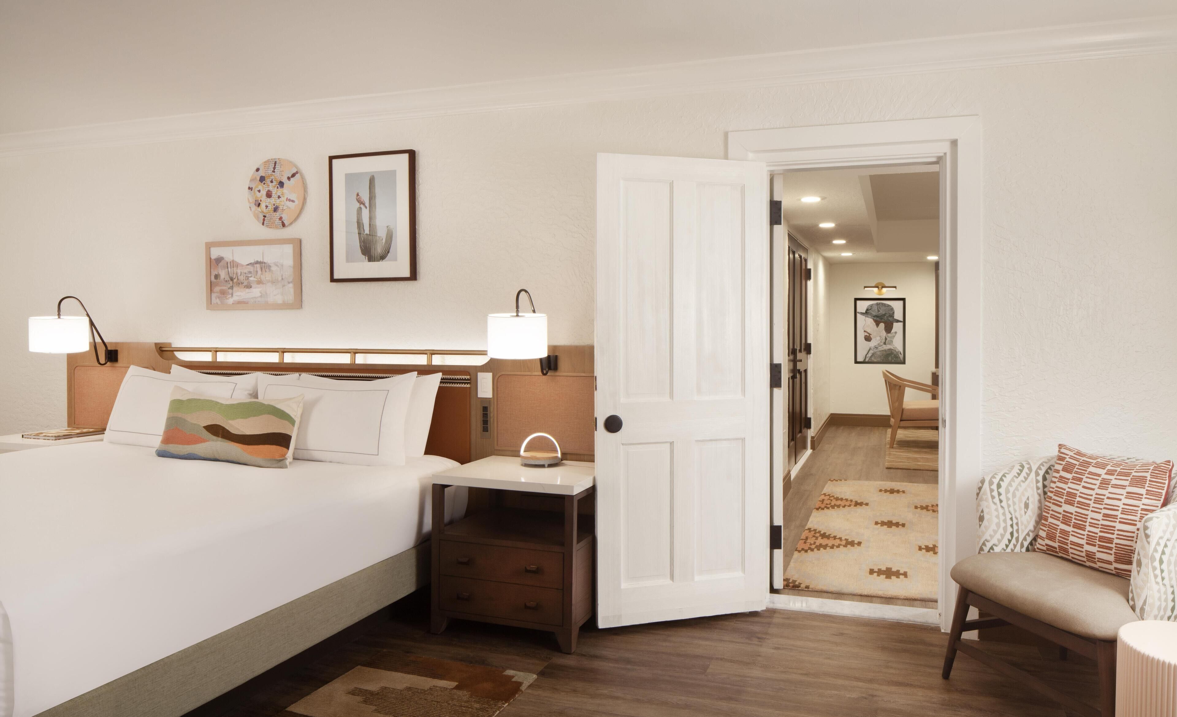 A hotel room with a bed, bedside tables, a nightstand, a chair, and a door leading to another room.