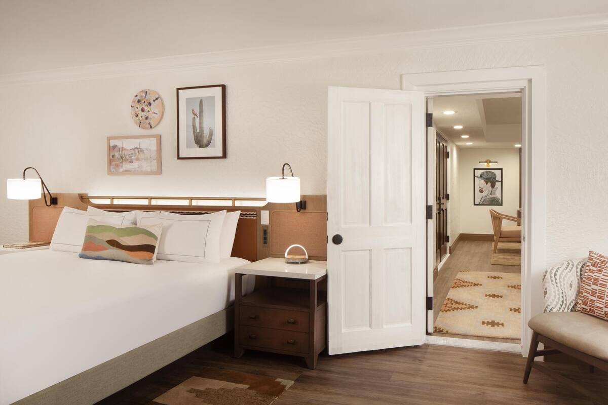 A hotel room with a bed, bedside tables, a nightstand, a chair, and a door leading to another room.