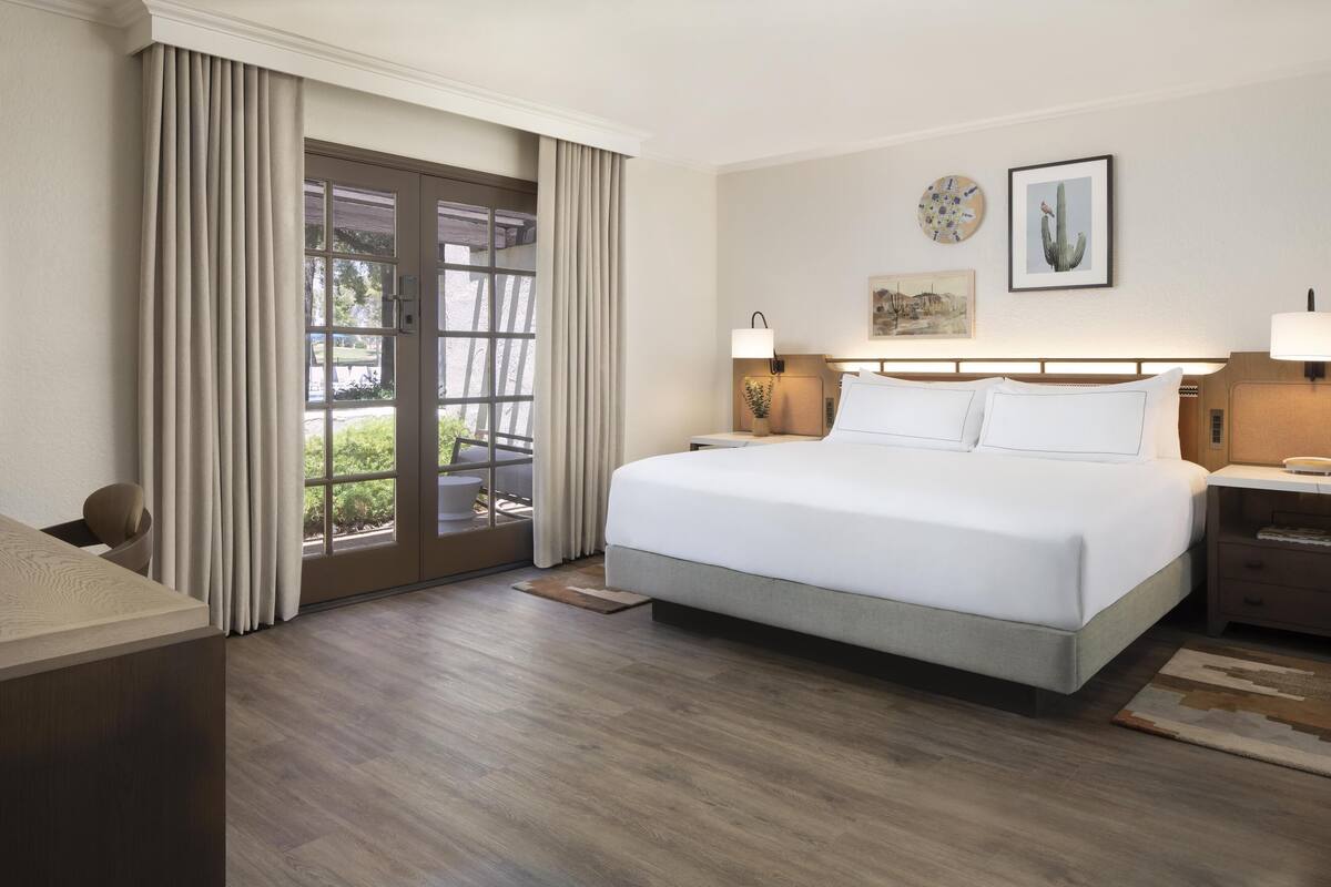 A hotel room with a large bed, a desk, a chair, a window with curtains, and a door leading to an outdoor area.