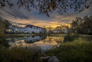 Vista del lago con un hotel, reflejado en el agua, rodeado de árboles y un cielo al atardecer.