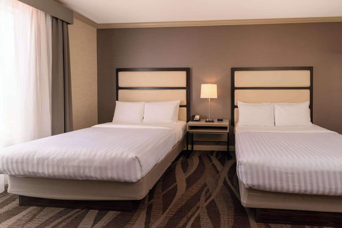 A hotel room with two beds, a nightstand, a lamp, and a large window with curtains.